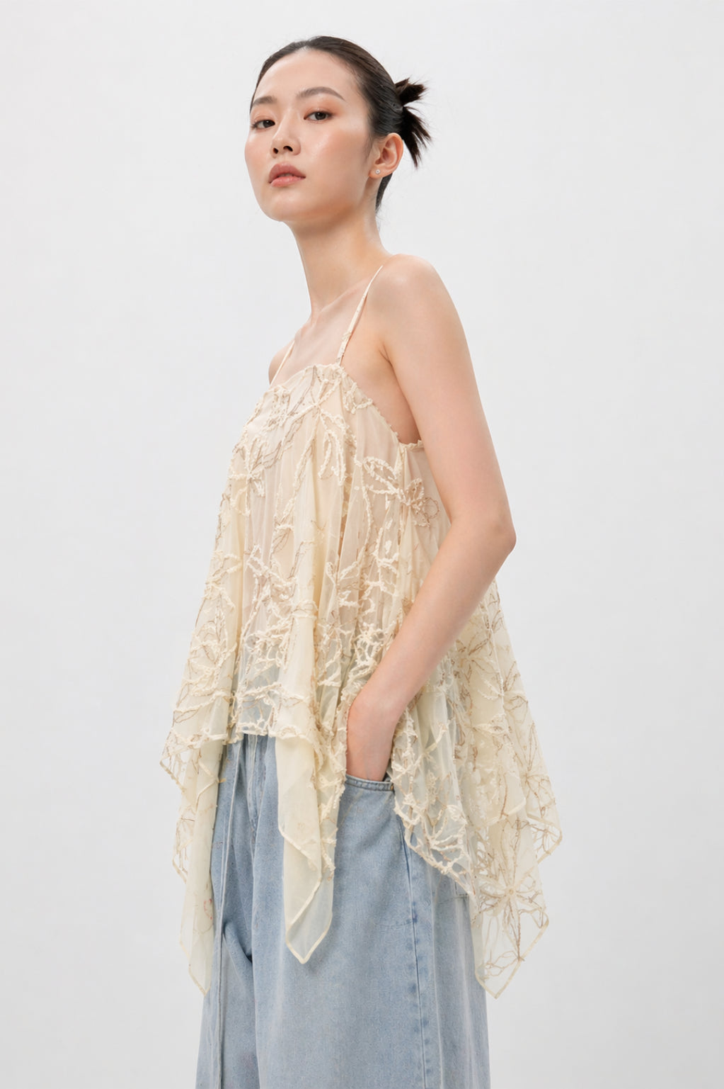Woman wearing a light-colored lace top and denim pants against a plain background