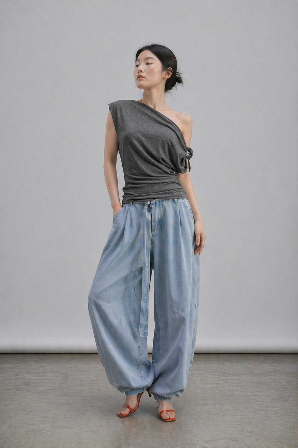 Woman wearing a gray sabrina top with buckle on one of the sleeve and light blue pants against a plain background