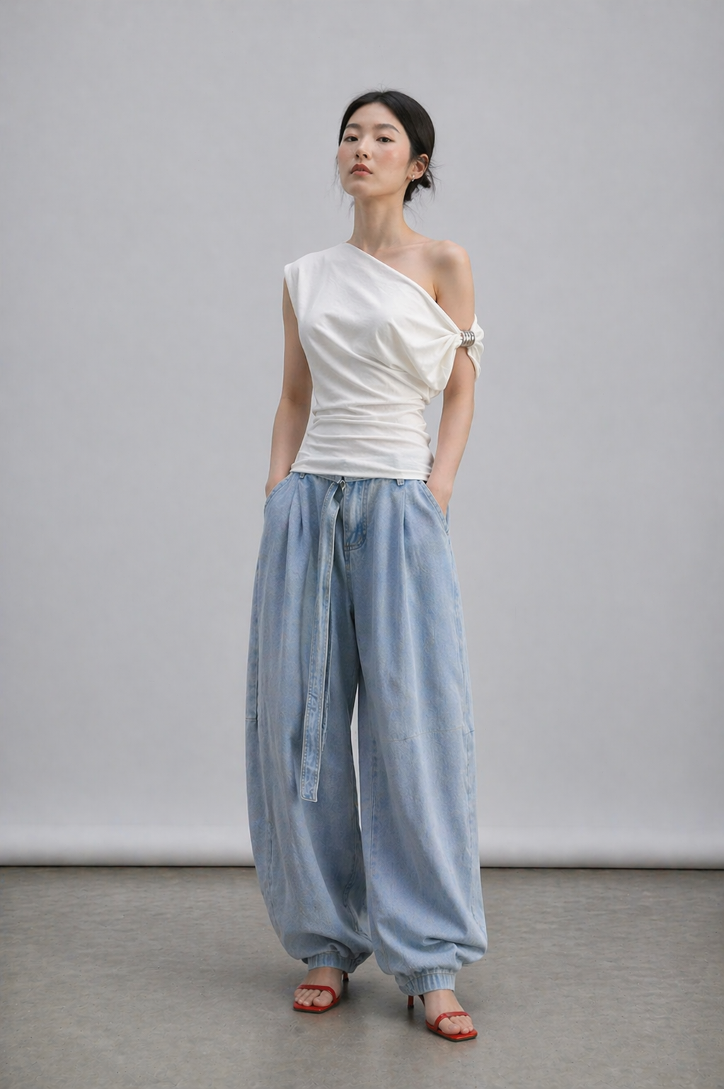 Woman wearing a white one-shoulder top and light blue pants against a gray background