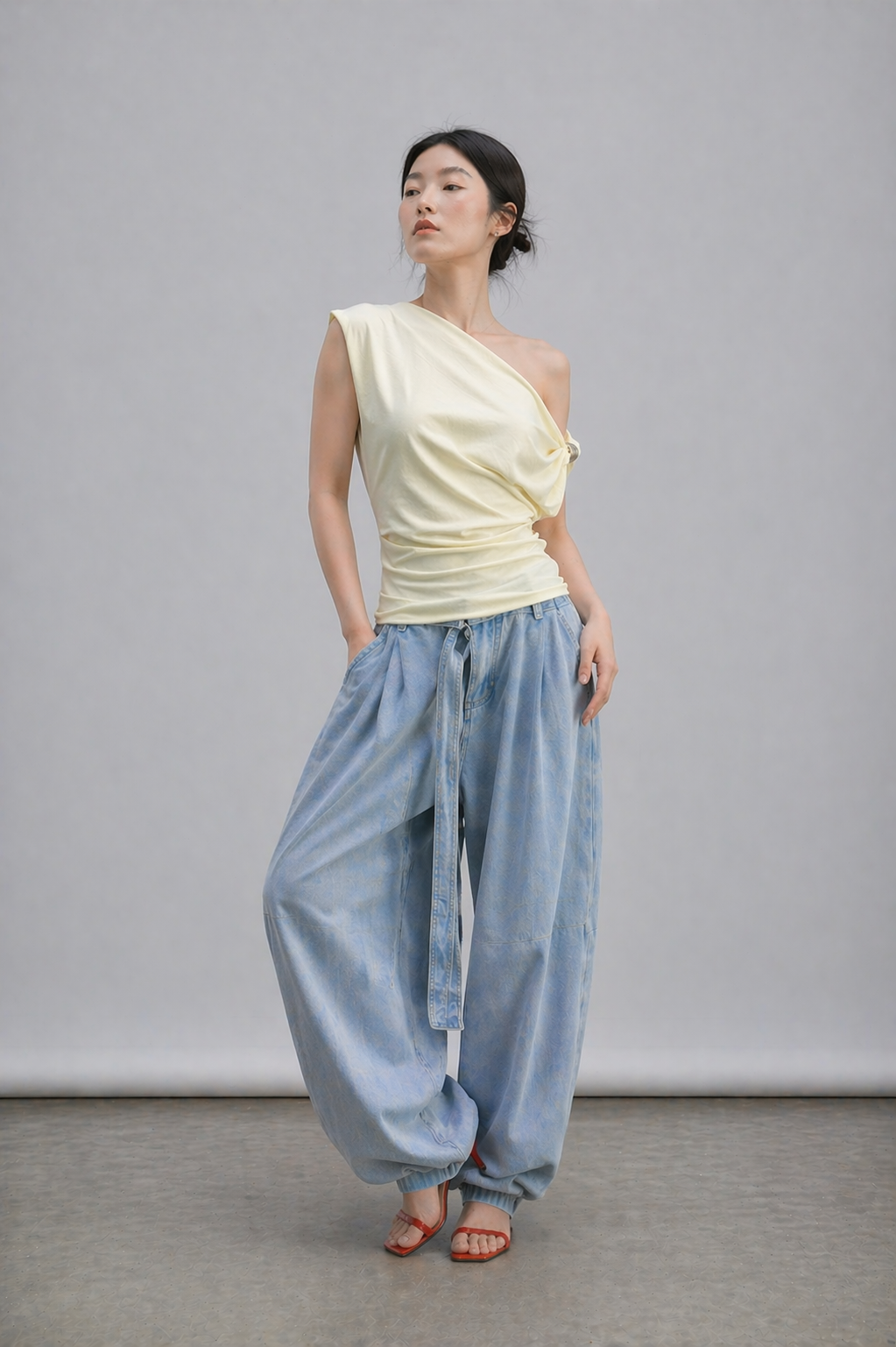 Woman wearing an off white one-shoulder top and blue high-waisted pants against a gray background
