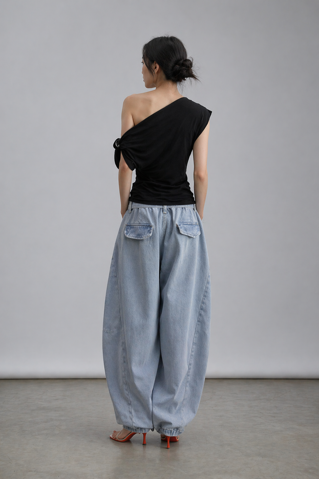 Woman wearing a black one-shoulder top and light blue denim pants against a plain background