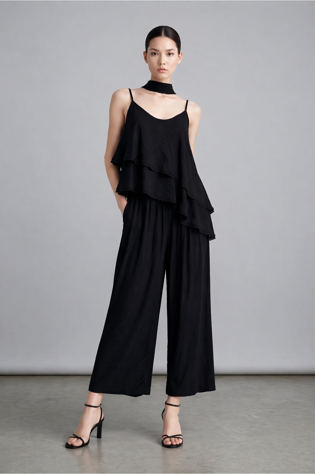 Woman wearing a black outfit with wide-leg pants and a layered top against a gray background