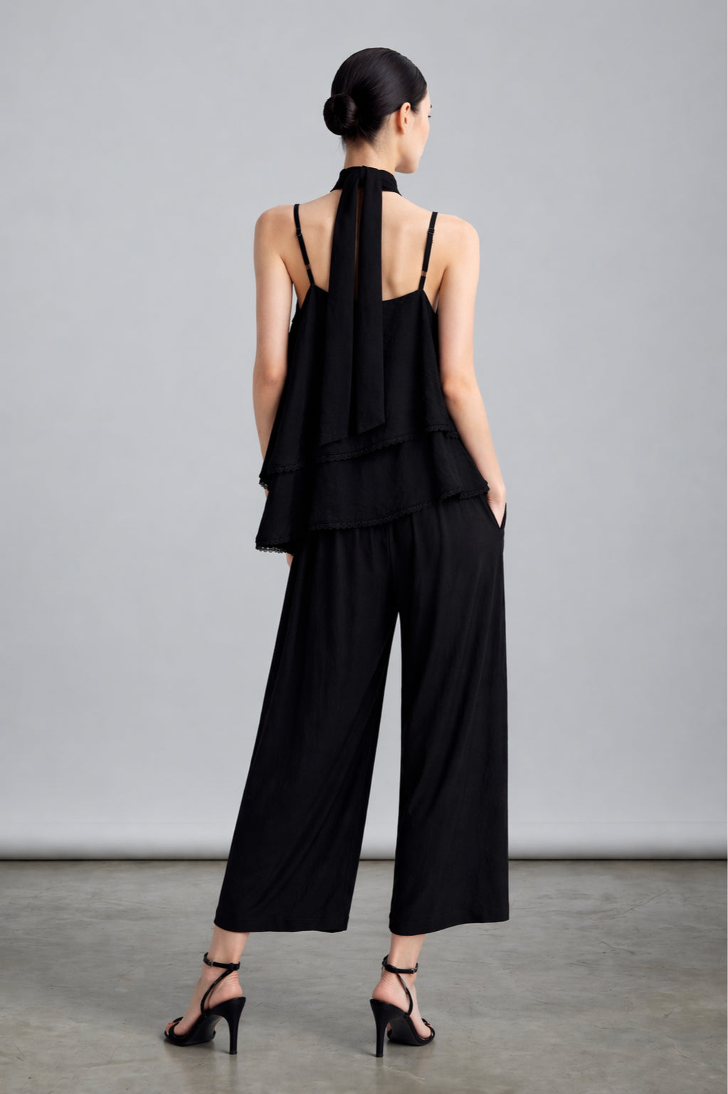 Woman wearing a black outfit with wide-leg pants from back against a gray background