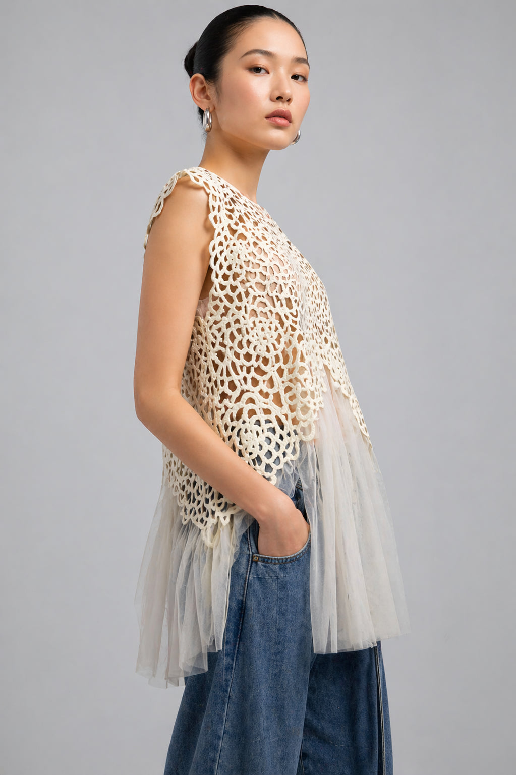 Woman wearing a sleeveless, patterned top with a sheer overlay and blue jeans on a gray background